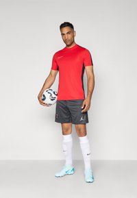 Punane Nike spordisärg mustade aktsentidega, hallitriibuline lühike püksid logoga, valged sokid ja helesinised jalgpalliketsid, hoiab mustrilist palli.