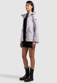 Femme portant une veste à capuche gris clair sur un short noir et des bottines noires, debout devant un fond clair uni.