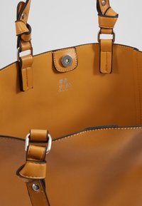 Kabelka z hnědé kůže s hladkým povrchem, stříbrnými kovovými prvky a černým kontrastním prošitím. Uvnitř je vyražené logo značky do materiálu.