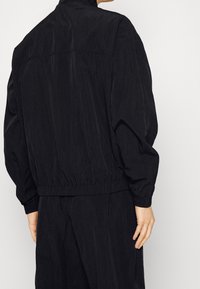 Chaqueta negra con una superficie texturizada, cintura y puños elásticos, alto cuello y un diseño simple y minimalista. Se muestra la vista posterior.