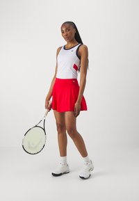 Tenniskläder som består av en vit linne med röda detaljer och en röd veckad kjol. Kombineras med vita sneakers och håller en tennisracket.