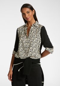 Schwarze und cremefarbene Bluse mit geometrischem Diamantmuster, langen schwarzen Ärmeln und einem Kragen. Verfügt über ein gebundenes schwarzes Kleidungsstück in der Taille.