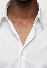 Chemise blanche à col ouvert, dotée d'un seul bouton au niveau du col. Le tissu semble lisse avec un léger éclat.