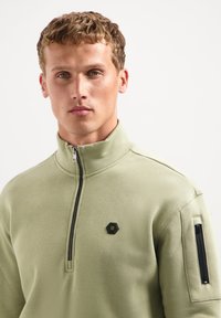 Lichtgroene sweater met een kwart ritssluiting en een hoge kraag, met een zwart hexagonaal logo op de borst en een ritssluiting zak op de mouw.