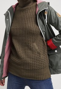 Chaqueta impermeable verde con un interior rojo y herrajes blancos, superpuesta sobre un suéter de punto de oliva texturizado. Ambos tienen un ajuste casual.