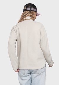 Hellbeige Fleecejacke mit strukturierten Oberflächen, hohem Kragen, langen Ärmeln und lockerer Passform, kombiniert mit einer schwarzen Mesh-Kappe.