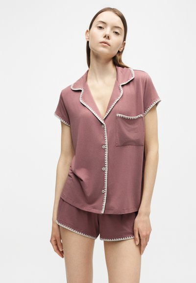 Jeune femme portant un haut boutonné à manches courtes rose poudré et un short assorti avec une bordure blanche festonnée, debout devant un fond clair uni.