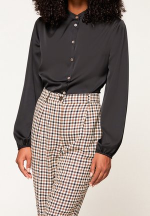 Vrouw draagt een zwarte blouse met lange mouwen en knopen, ingestopt in een broek met hoge taille in bruin, zwart en wit geruit patroon.