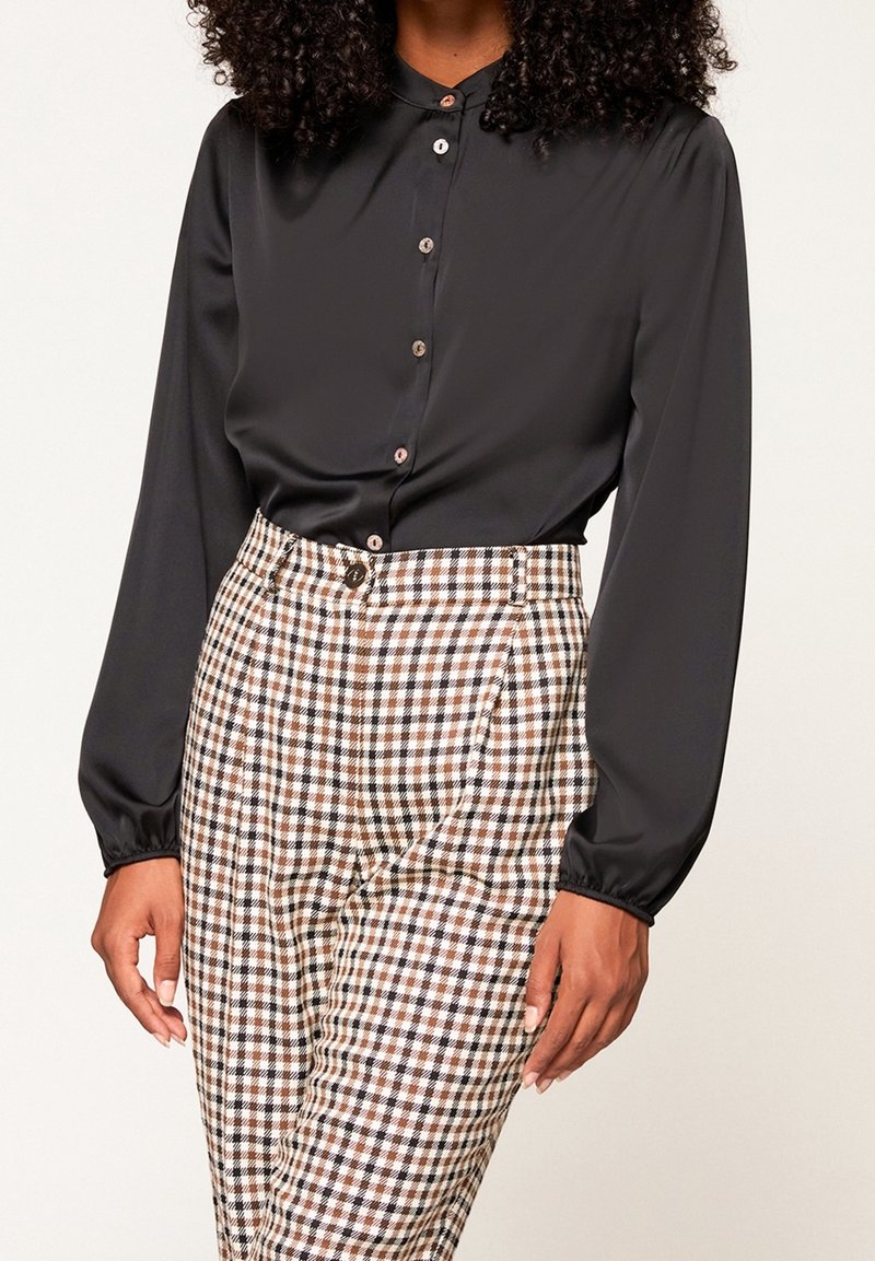 Femme portant un chemisier noir à manches longues boutonné, rentré dans un pantalon à taille haute à carreaux marron, noir et blanc.