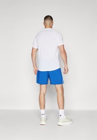 Camiseta atlética blanca de mangas cortas, combinada con pantalones cortos azules. La tela parece ligera, ideal para el deporte. Logos destacados visibles.