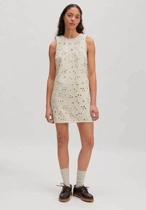 Vrouw draagt een mouwloze witte mini-jurk met oogjes, lichtgekleurde sokken en zwarte veterschoenen, staand tegen een eenvoudige lichte achtergrond.
