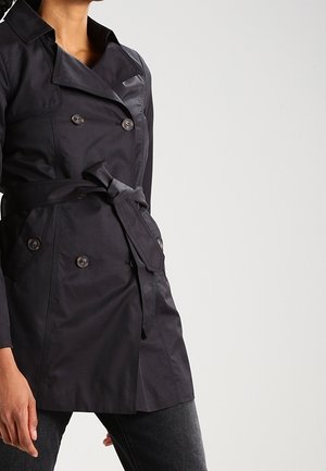 Femme portant un trench-coat noir croisé ceinturé avec des poches à boutons, associé à un pantalon foncé sur un fond uni.