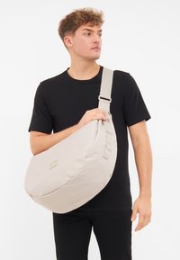 Borsa a tracolla beige realizzata in tessuto morbido, con forma arrotondata, tracolla regolabile e un piccolo tag con logo frontale. Texture liscia, design minimalista.