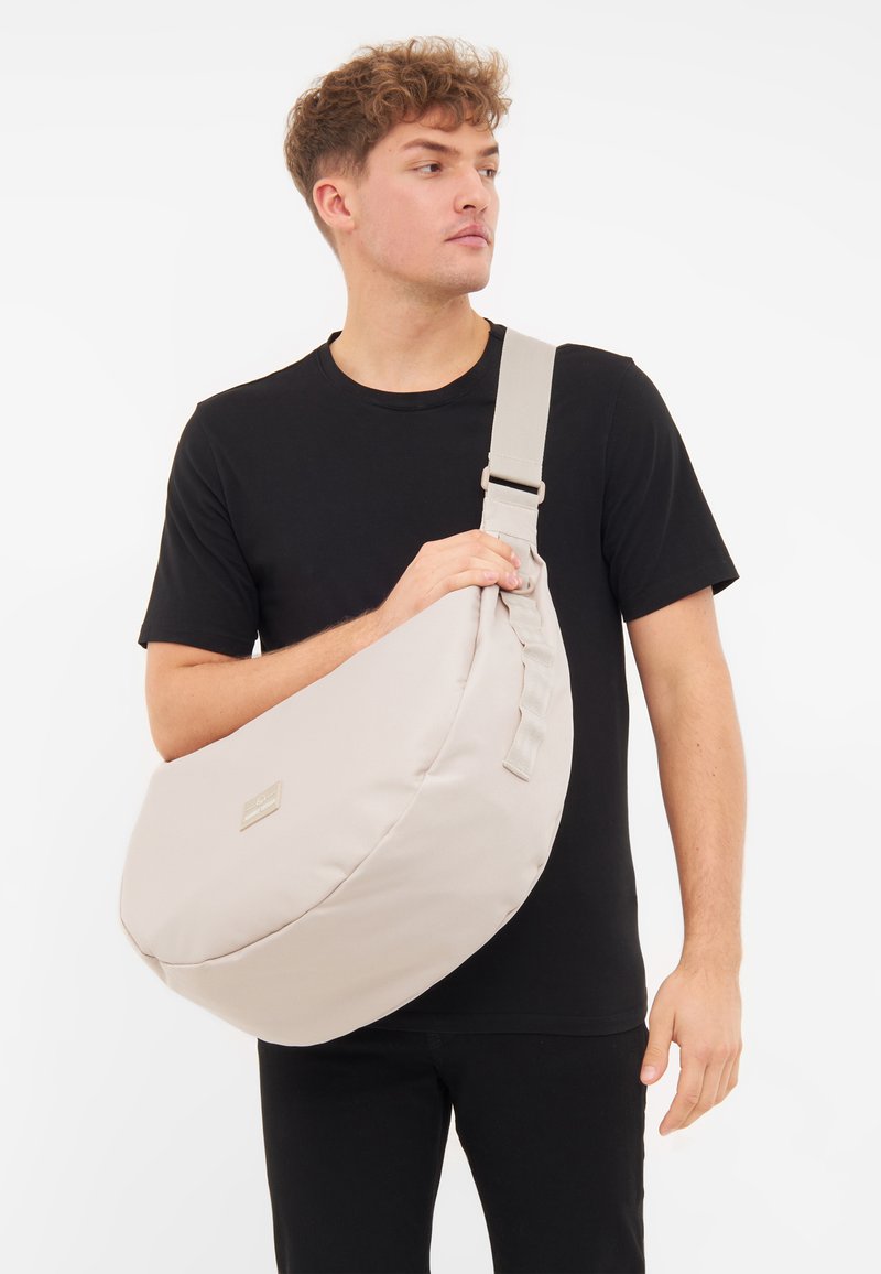 Borsa a tracolla beige realizzata in tessuto morbido, con forma arrotondata, tracolla regolabile e un piccolo tag con logo frontale. Texture liscia, design minimalista.