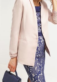 Ljust rosa blazer i mjukt material, bärs över en marinblå klänning i spets. Outfiten kompletteras av en liten marinblå handväska.