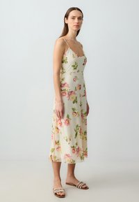 Femme debout dans une robe crème mi-longue à imprimé floral rose et vert, portant des sandales plates blanches à lanières sur un fond léger uni.