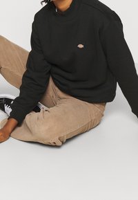 Sudadera negra con cuello redondo y pequeño logo, combinada con pantalones de pana beige. El modelo se sienta sobre una superficie plana, vistiendo zapatillas negras.