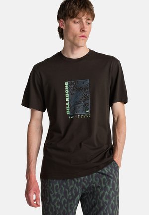Młody mężczyzna w czarnej koszulce Billabong z nadrukiem mapy topograficznej i napisem „Adventure Division”, w połączeniu z ciemnozielonymi spodniami w wzory.