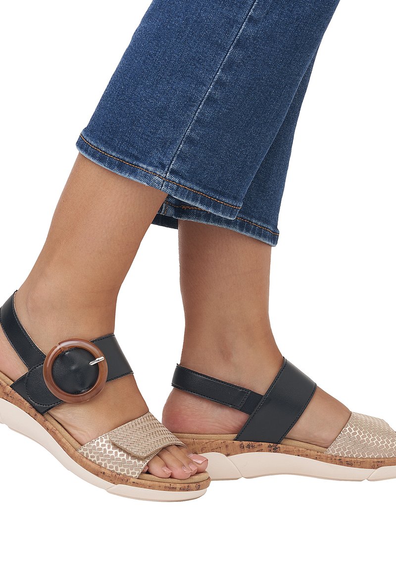 Due sandali: uno presenta una tomaia intrecciata beige con suola in sughero; l'altro ha una cinghia nera e un accento circolare marrone.