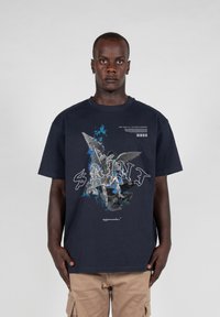 Marineblå grafisk t-shirt med et detaljeret print af en engel og en figur, med ordet "SAINT" i hvide stiliserede bogstaver. Lavet af bomuldsmateriale.