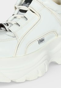 Sneakers bianche del marchio Buffalo con suola spessa, cuciture dettagliate e lacci arrotondati, visualizzate in primo piano di lato.