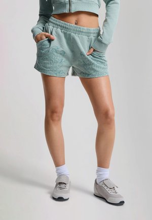 Mujer con pantalones cortos de estampado floral en azul claro, sudadera corta a juego, calcetines blancos y zapatillas beige, de pie con las manos en los bolsillos.