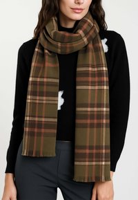 Donna che indossa una sciarpa a quadri verde oliva e marrone sopra un maglione nero e pantaloni scuri, in piedi contro uno sfondo semplice.