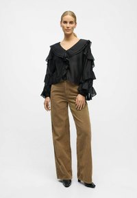 Une femme se tient debout, portant un blouse noire à volants, un pantalon en velours côtelé large marron et des chaussures noires à talons pointus, sur un fond blanc uni.