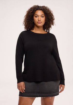 Femme aux cheveux bouclés portant un pull noir à manches longues et une jupe en jean gris foncé, debout devant un fond uni.
