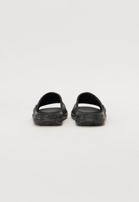Fekete, nyitott orrú, slip-on szandálpár mintás talppal, sima fehér háttér előtt.