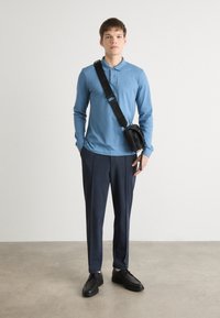 Helles blaues Langarm-Polohemd, dunkle maßgeschneiderte Hose, schwarze Schuhe und eine kleine schwarze Umhängetasche. Glatter Stoff, taillierte Passform.