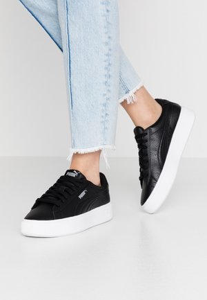 Osoba ubrana w jasnoniebieskie dżinsy z postrzępionymi nogawkami i czarne sneakersy Puma z białymi podeszwami, stojąca na jednolitej białej podłodze.