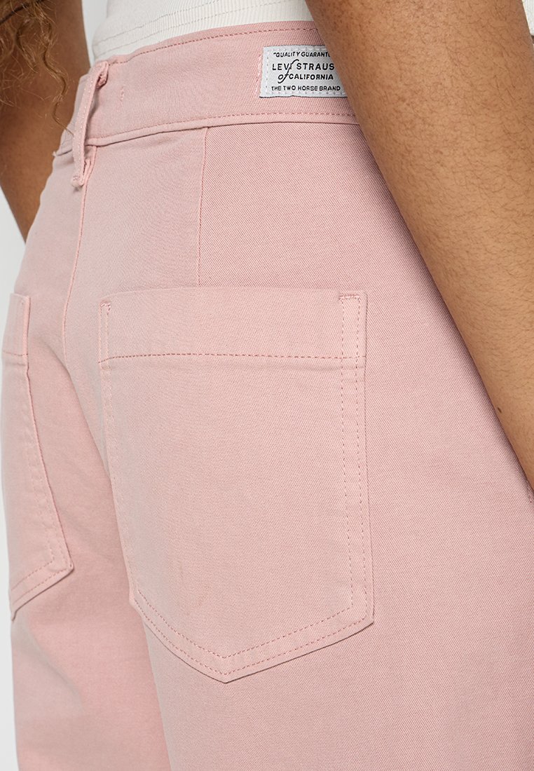 Les pantalons en denim rose présentent de doubles poches arrière et des détails de couture subtils. Le tissu a une texture lisse avec une coupe décontractée et une fermeture éclair.