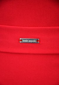 Czerwony materiał o gładkiej fakturze ma srebrną prostokątną metkę wygrawerowaną z napisem "Kasia Zaporza", przymocowaną do jednego z brzegów.