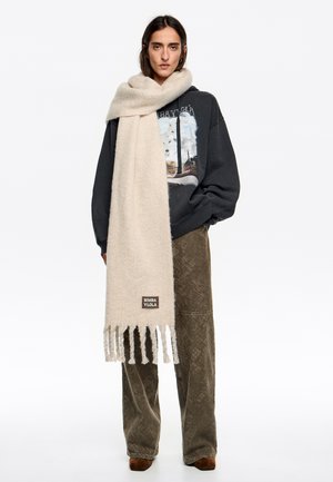 Bufanda de lana beige con bordes deshilachados y un parche de logo, sobre un suéter oscuro, combinada con pantalones marrones texturizados.