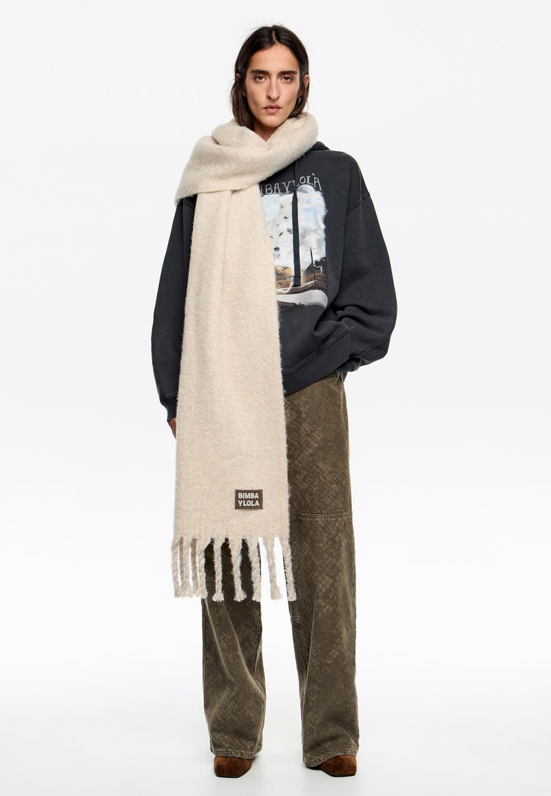 Bufanda de lana beige con bordes deshilachados y un parche de logo, sobre un suéter oscuro, combinada con pantalones marrones texturizados.