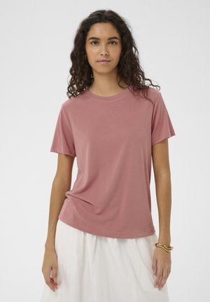 Mladá žena s kučeravými tmavými vlasmi nosí prachovo ružové tričko s krátkym rukávom a bielu sukňu, stojaca na jednoduchom svetlom pozadí.