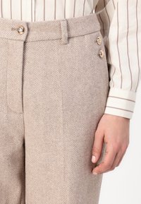 Pantalons en chevron beige à taille haute, avec une fermeture à bouton et deux boutons décoratifs sur le côté. Une texture lisse est visible.