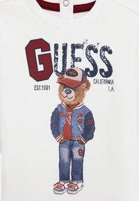 Weißes T-Shirt mit einem Grafikdruck eines Teddybären in einer blauen Jacke, rotem Shirt und Mütze, mit dem Text "GUESS" und zusätzlichen Markeninformationen.