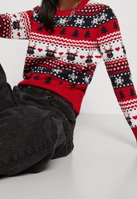 Roter und schwarzer Pullover mit Muster aus Rentieren, Herzen und Bäumen. Strickdesign mit gerippten Bündchen und Saum. Kombiniert mit dunklen Jeans.