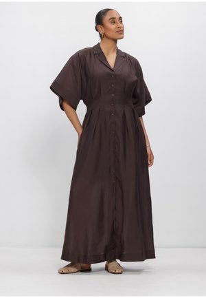 Frau trägt ein dunkelbraunes, knöpfbares Maxikleid mit kurzen Ärmeln, Seitentaschen und beigen flachen Sandalen, steht vor einem schlichten Hintergrund.