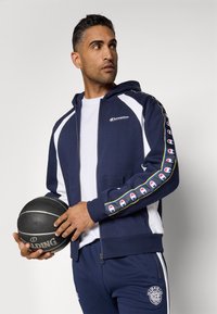 Sweatshirt azul-marinho com fecho e painéis brancos, apresentando um logótipo e riscas multicoloridas nas mangas. Calças de treino azul-marinho combinando e uma bola de basquete.