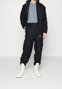 Svart ytterjacka med hög krage, grå ribbad tröja, cargobyxor med fickor och vita snörade stövlar.