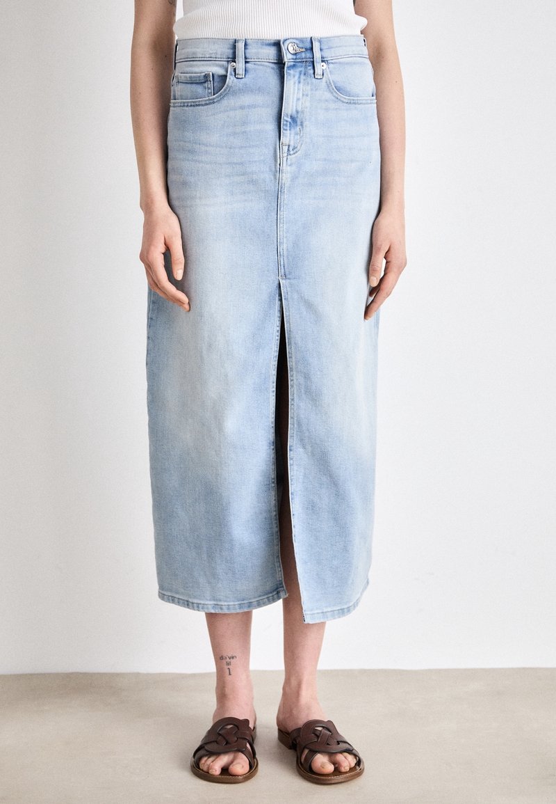 Femme portant une jupe midi en jean bleu clair avec une fente à l'avant, un top blanc côtelé, des sandales marron et un tatouage à la cheville, debout sur un sol beige contre un mur blanc.