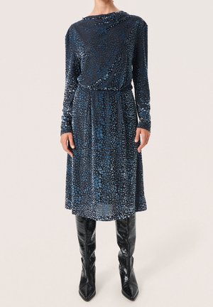 Kobieta ubrana w niebieską, wzorzystą sukienkę midi z długim rękawem, w połączeniu z czarnymi, teksturowanymi skórzanymi kozakami za kolano.