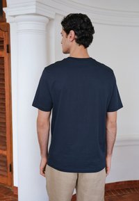 Homme aux cheveux bouclés et foncés debout à l'intérieur, faisant face à un mur blanc, portant un t-shirt bleu marine et un pantalon beige.