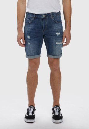 Uomo che indossa pantaloncini di jeans blu consumati risvoltati, t-shirt bianca e scarpe da ginnastica nere e bianche con lacci, in piedi su sfondo bianco.