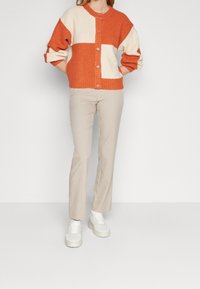 Cardigan en tricot coloré en orange et crème avec un col rond et une fermeture à boutons. Associé à un pantalon beige et des baskets blanches.
