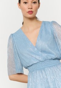 Jasnoniebieska sukienka z dekoltem w serek, krótkimi przezroczystymi rękawami i marszczoną talią. Materiał ma subtelną, błyszczącą teksturę i pionowe paski.