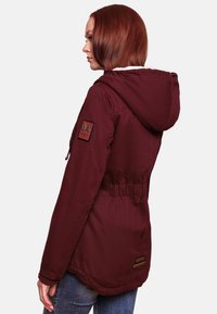 Burgundy parka jacket with a fur-lined hood, elastic waist, and logo patch on the sleeve. Featuring a smooth, durable fabric and side pockets.